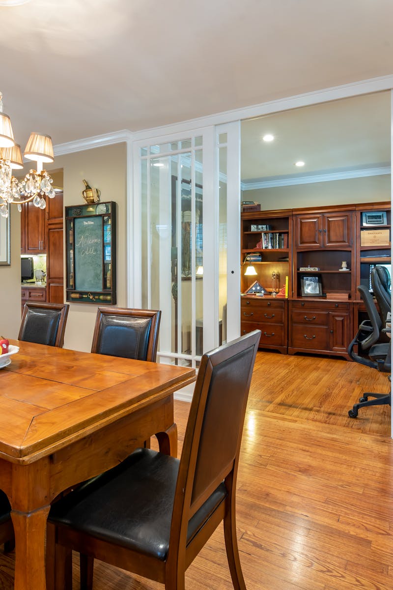 A cozy dining room with a wooden table, leather chairs, and a chandelier, connected to an office space.